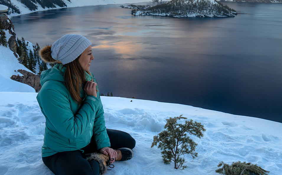 A woman sitting on the snowy ground near a lake.