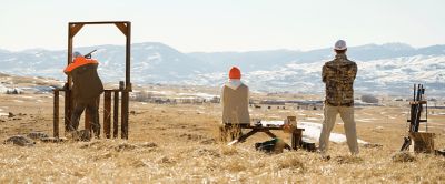 A hunter in an orange Columbia Sportswear parka shoots at a target as two fellow hunters look on. 