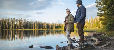 Cozied up in the Women's Super/DS™ Stretchdown Parka and the Men's Super/DS™ Stretchdown Climb Shacket, at camp near a lake in the trees.