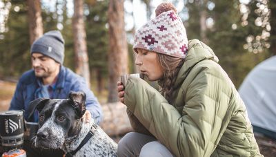 At the camp fire, warming up with a cup of coffee and pup by her side, wearing the Women's Super/DS™ Stretchdown Parka