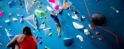Jesse climbing in a gym, Kyra Condie sitting watching.