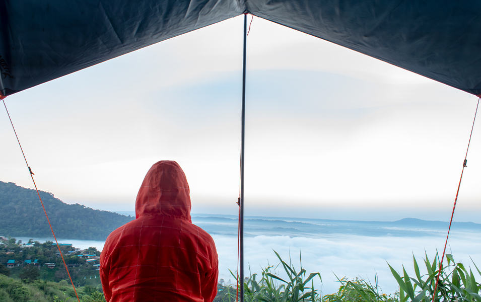 A camper in a red Columbia Sportswear rain jacket looks out on a wet day from beneath a camping tarp.