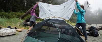Two women hang a tarp over a tent while camping in the rain.