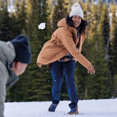 A woman throwing a snowball in a snowy forest bundled up in a jacket.