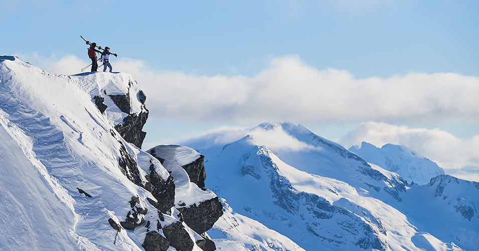 Two skiers standing on top of a high mountain cliff.
