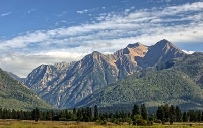 Beyond a grass-covered meadow, steep, tree-covered slopes flank high alpine peaks.