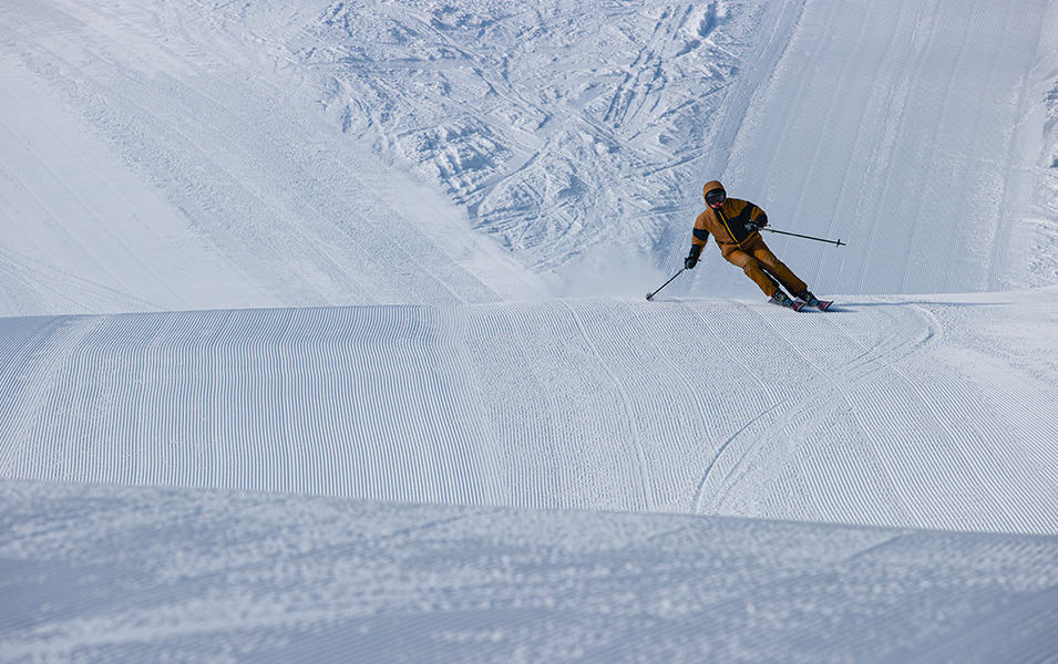 A man in an orange jacket skis down a patch of corduroy on a freshly groomed ski slope. 