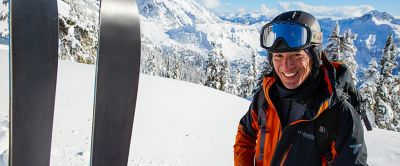 A skier in a red Columbia Sportswear jacket smiles at the camera on a beautiful powder-filled mountain as his skis stick out of the snow in the foreground.  