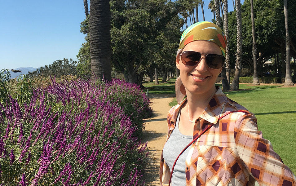 A woman in a plaid shirt and headscarf smiles at the camera on a sunny day at Santa Monica’s Palisades Park.
