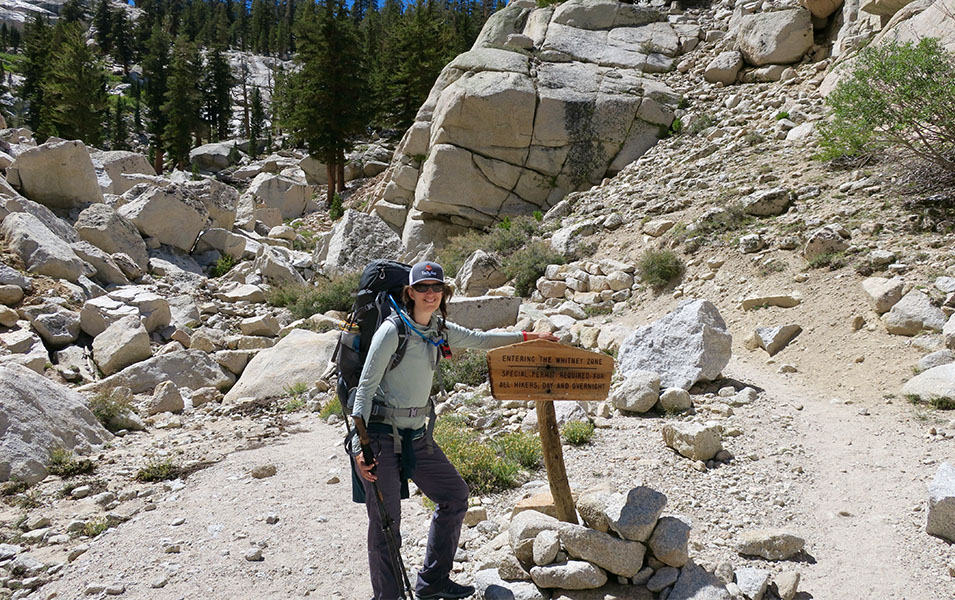 A woman stands at the base of the Mt. Whitney trail carrying a backpack and smiling for the camera. 
