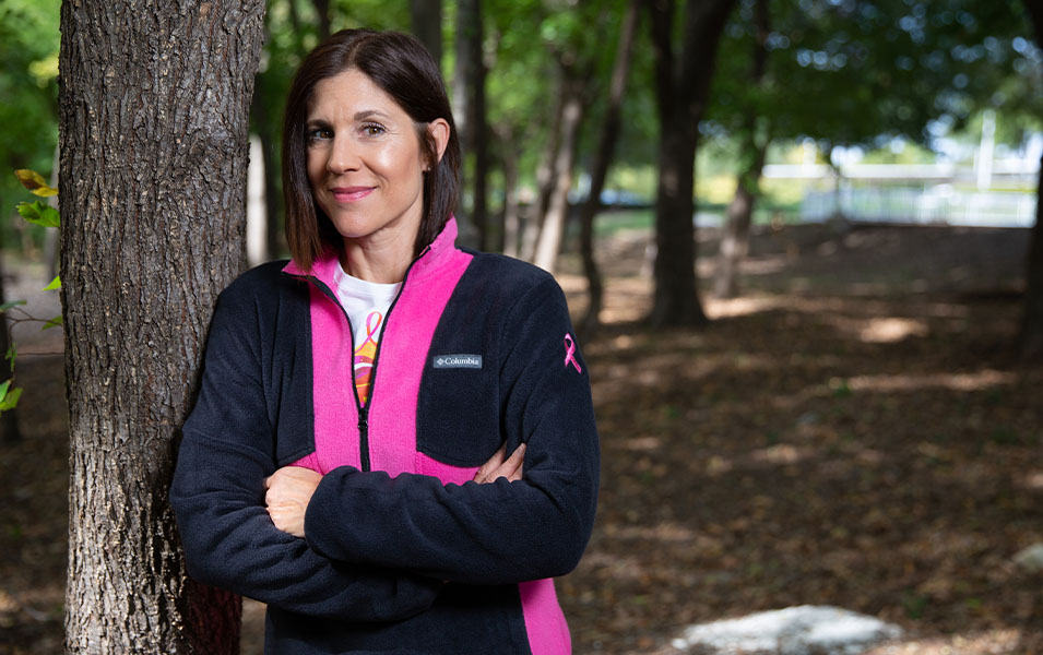 A woman wearing a black and pink fleece jacket leans against a tree and looks at the camera.