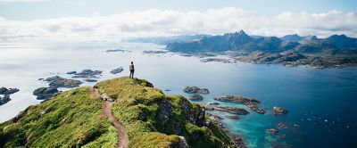 A hiker in search of friluftsliv stands on a lush green hilltop in Norway staring out into the ocean.