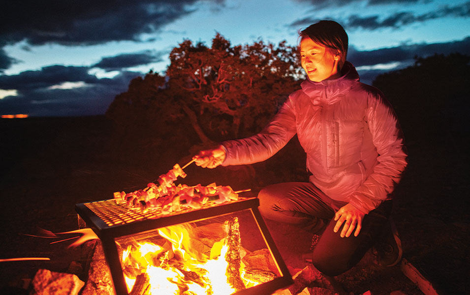 A woman puts a log on a fire in a nighttime wilderness setting.