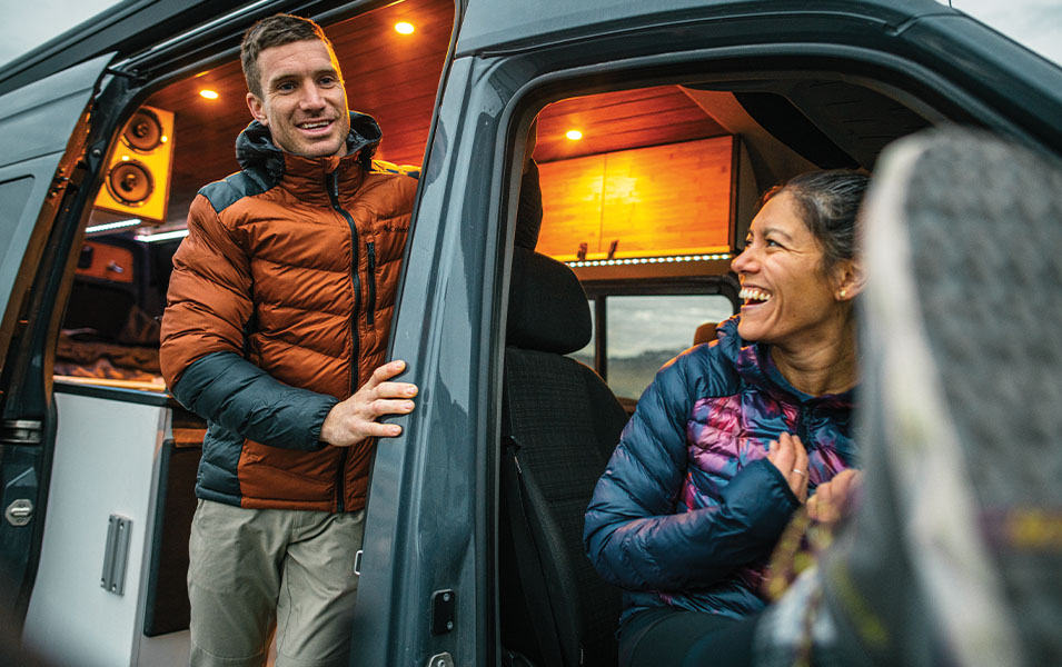 A man in an orange Columbia Sportswear jacket leans out of a van as a woman laughs in the front seat. 