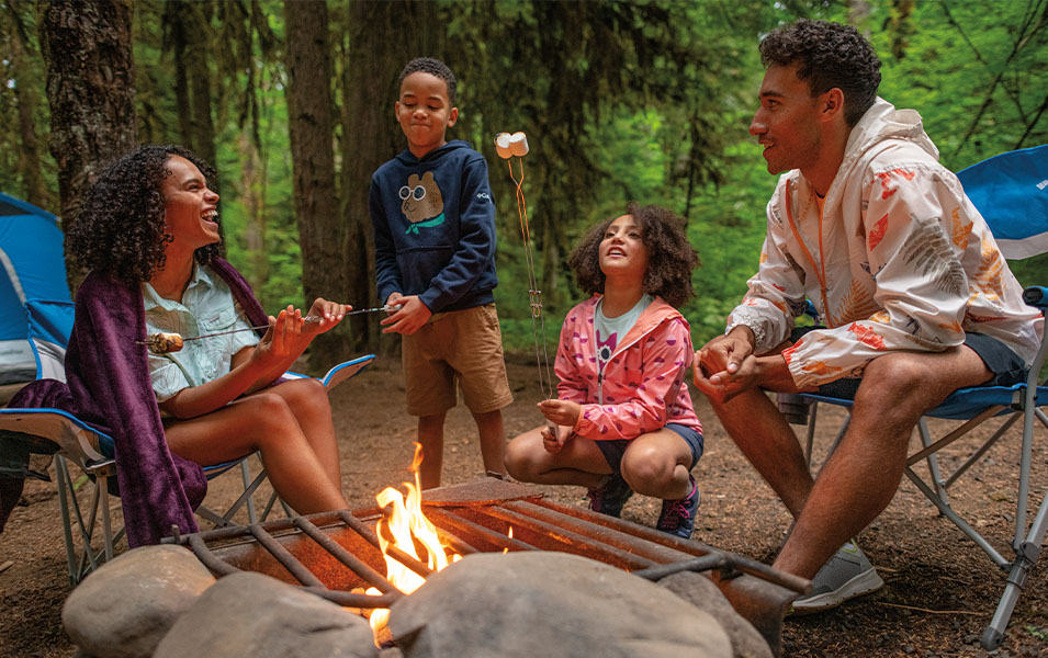A family roasts marshmallows over a campfire in a pretty wooded area. 