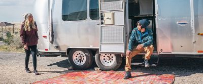 A woman walks toward a man sitting on the steps of a retro-looking RV.