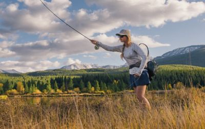 A woman on a grassy river bank demonstrates perfect fly casting technique.