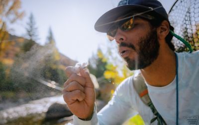 A man wearing sunglasses and a ball cap looks closely at a white fly. In the background is his fishing net.