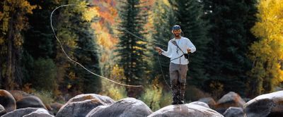 Man wearing a brimmed cap uses a fly rod to cast a line into a creek.