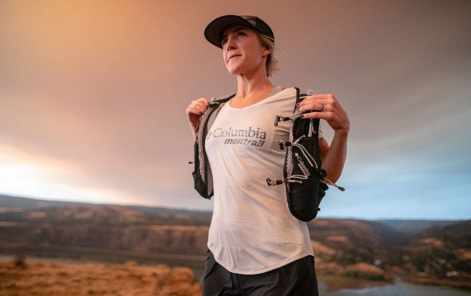 A woman in a white Columbia Sportswear shirt and black hydration vest pauses to cool down after a trail run.