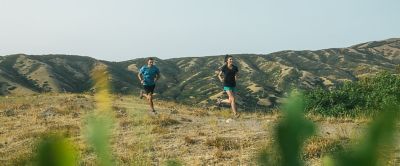 Two trail runners jog along a scenic high desert trail as the sun shines down overhead. 