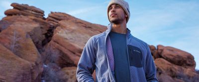 A man stands in front of a scenic outdoor setting wearing a blue Columbia Sportswear midlayer with Omni-Heat™ Helix technology. 