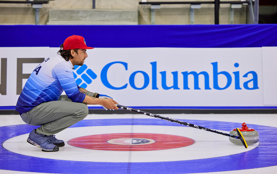 Man crouching on the ice with curling rock and a broom. 