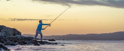 A man with a long fishing pole casts his line out into the water. The sky is turning orange and the water is choppy.