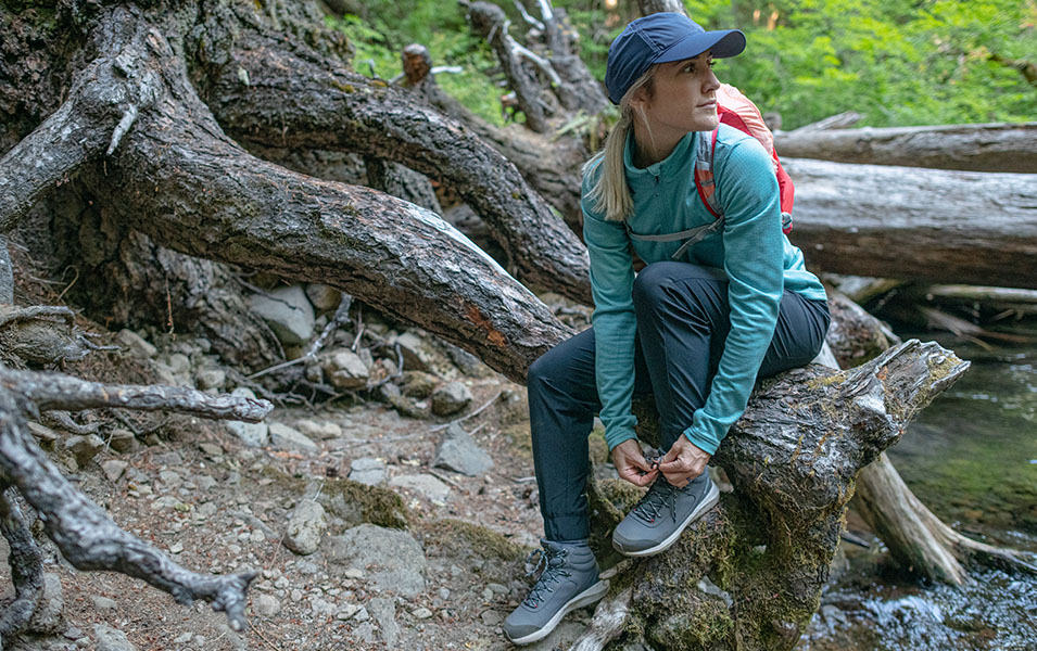 A woman wearing a blue Columbia Sportswear shirt sits on a log in the forest tying the laces of her hiking shoes.