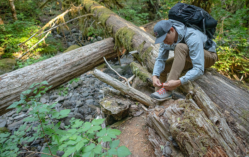 A man wearing a light blue Columbia Sportswear jacket stoops down on a mossy log in the woods to lace his hiking boots. 
