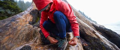 A women wearing a red Columbia Sportswear jacket stoops down to tie the laces of his hiking boots. 