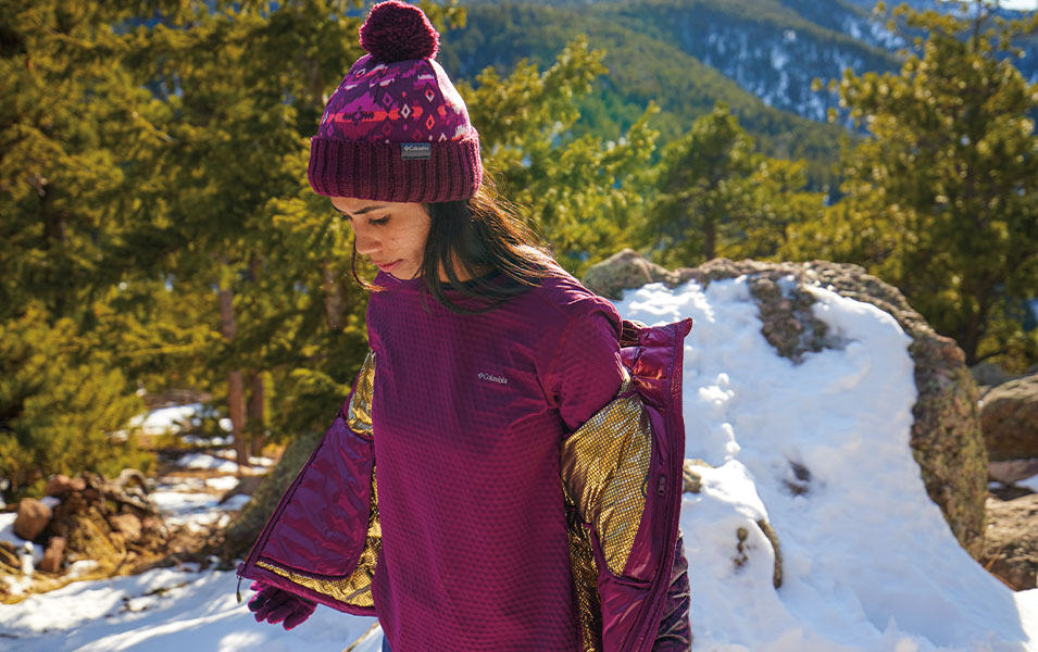 A woman in a bright purple Columbia Sportswear jacket and hat takes her jacket off in front of a scenic mountain trail.  

 