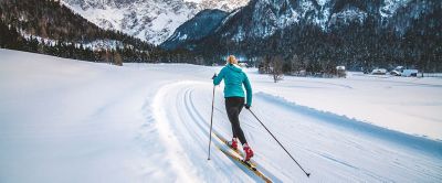 A woman in a turquoise top and black pants cross country skis away from the camera toward a beautiful mountain landscape