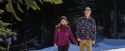 A pair of hikers walk through a snowy field wearing fleece jackets. 