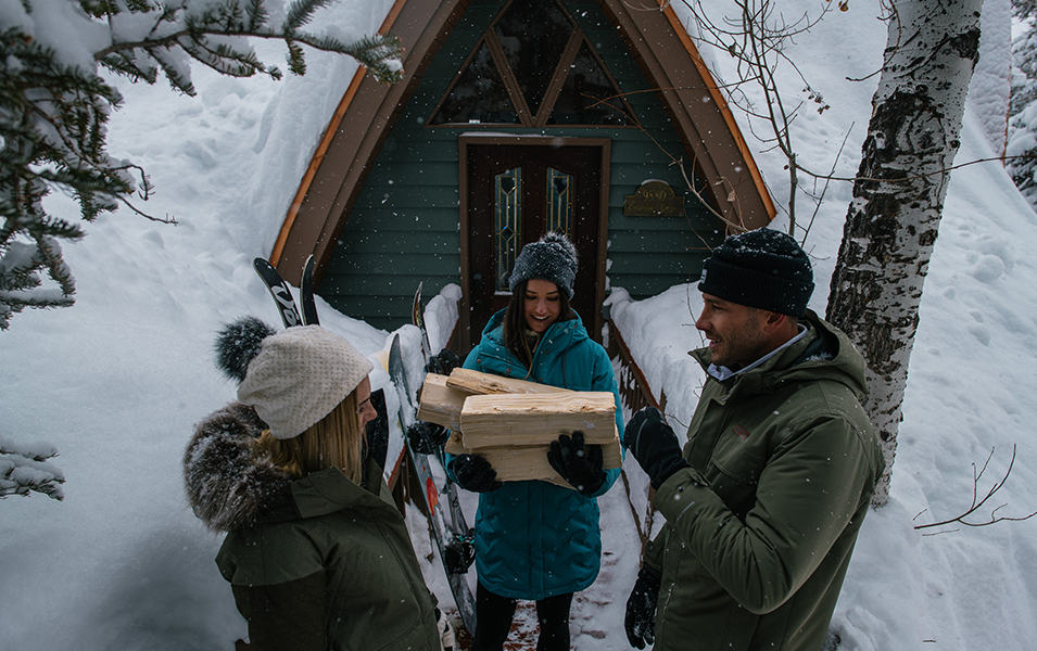 A group of friends stands outside a winter cabin chatting while one holds firewood. 