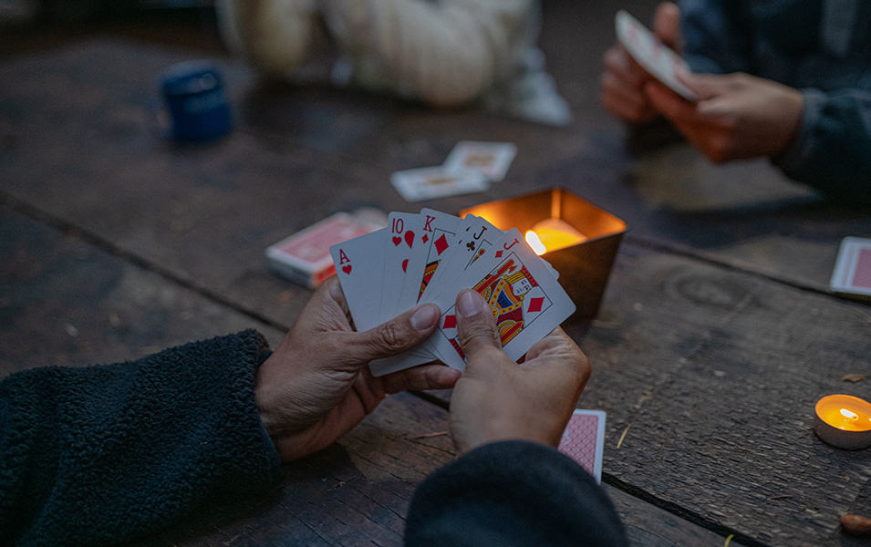 A close-up shot of someone’s hands holding playing cards at a rustic picnic table set with lighted candles. 