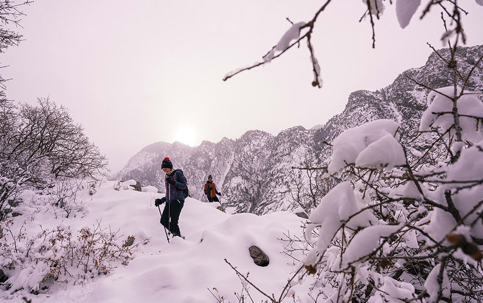 A woman treks through a snowy winter wonderland with another hiker and a stretch of mountains in the background. 