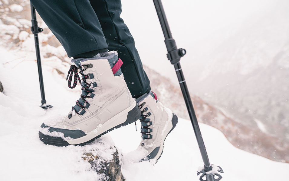 A close-up photo of a pair of white winter hiking boots and trekking poles as they climb up a snowy mountain trail. 