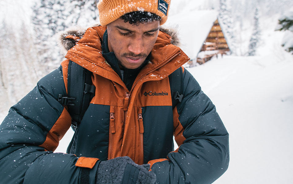 A man in an orange parka and beanie adjusts his gloves amid a scenic winter setting.  