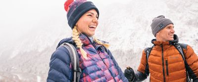 A man in an orange Columbia Sportswear puffer jacket laughs with a woman wearing a purple jacket and winter beanie in a snowy mountain landscape.