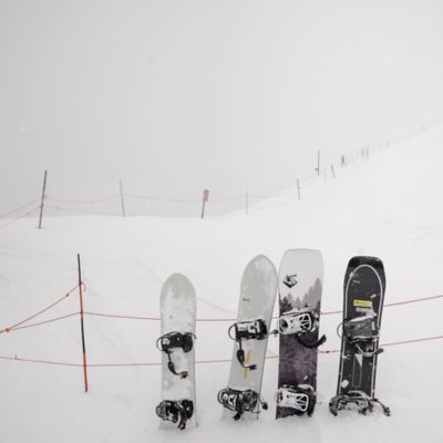 Snowboards lined up in the snow leaning against a fence.