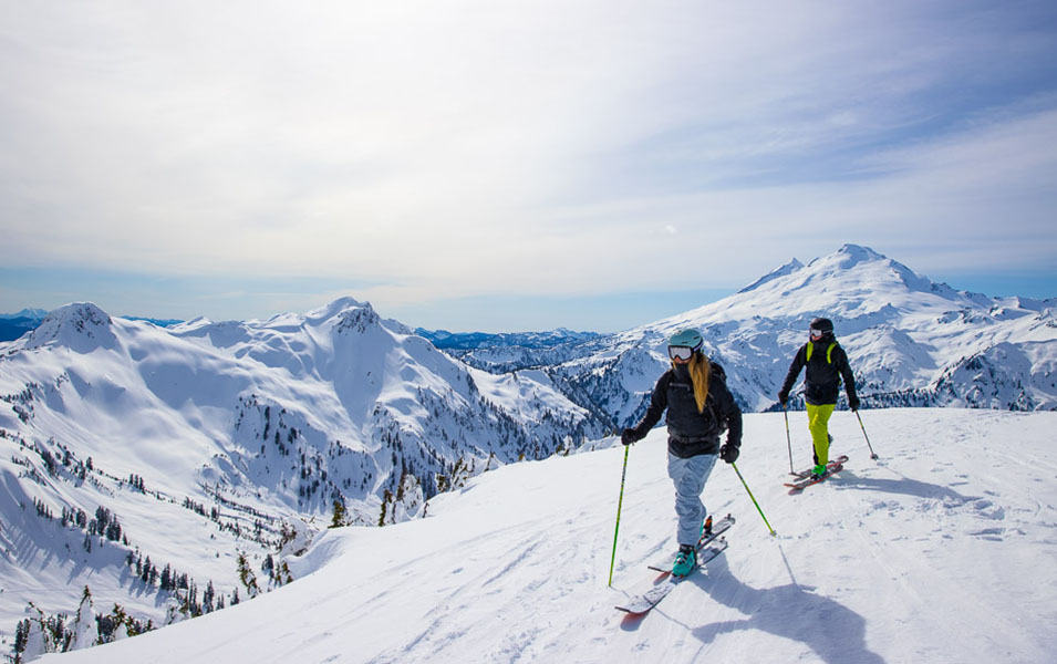 Two skiers stand on top of a gorgeous mountaintop on a sunny day with a sweeping panoramic view behind them. 