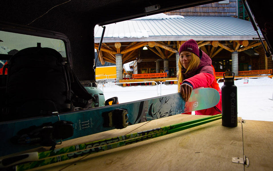A woman in a reddish pin jacket pulls a set of skis out of her car with a ski lodge in the background. 