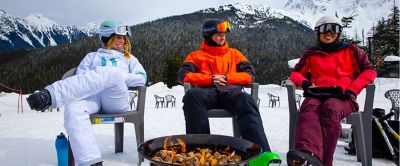 Three skiers sit around a firepit at a ski resort smiling with mountains in the background.  