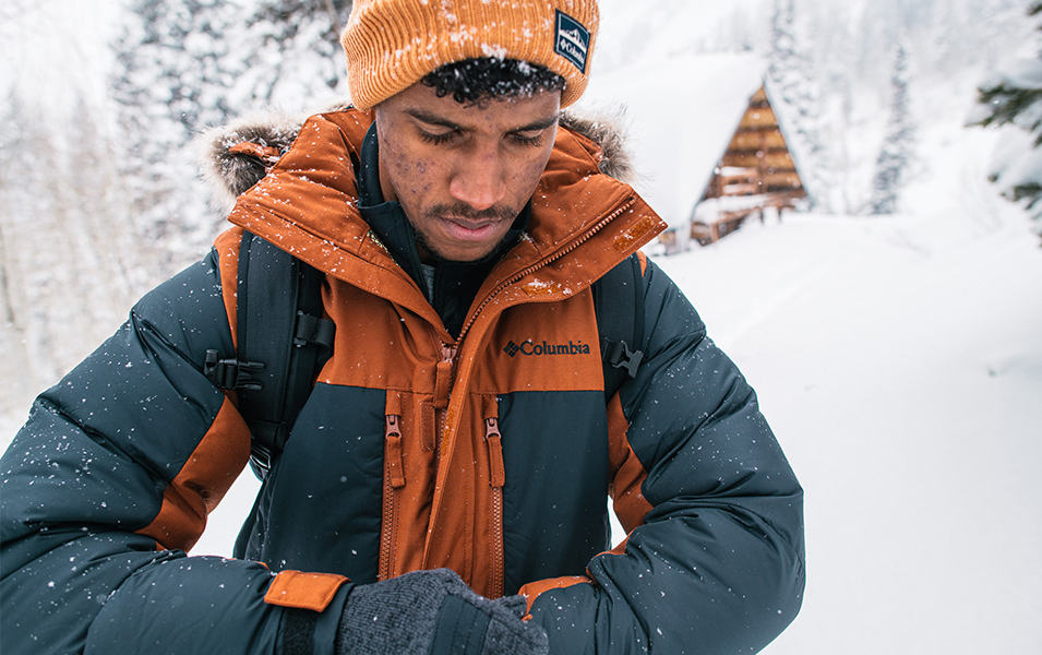A man stands in the snow looking down wearing a gray-and-orange Columbia Sportswear insulated jacket.