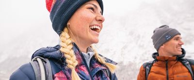 A woman with blonde braids wearing a winter hat stands in the snow as a man wearing an orange insulated jacket smiles in the background.