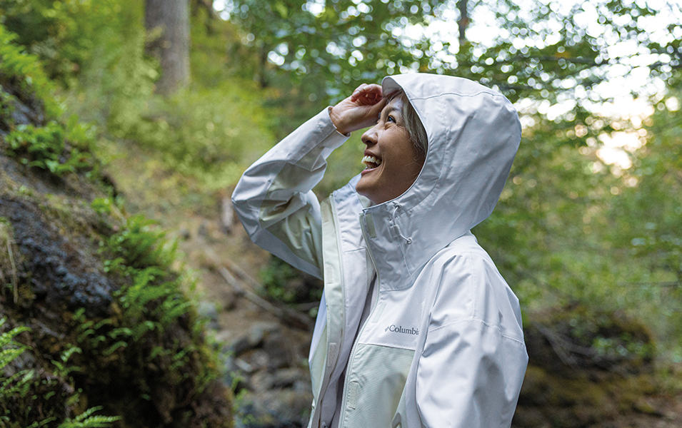 A woman stands on a hiking trail wearing a white Columbia Sportswear Omni-Tech™ jacket and laughing.