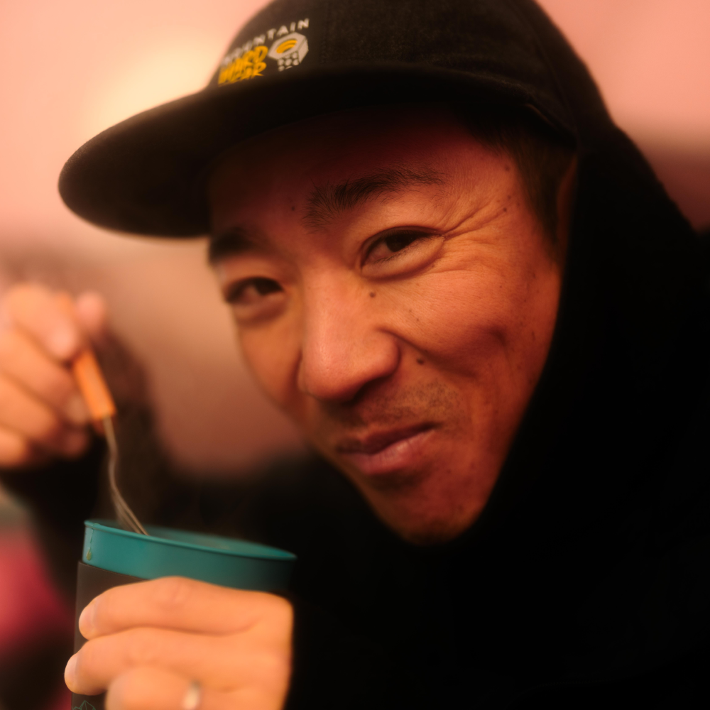 Daisuke smiles at the camera while eating ramen at basecamp.