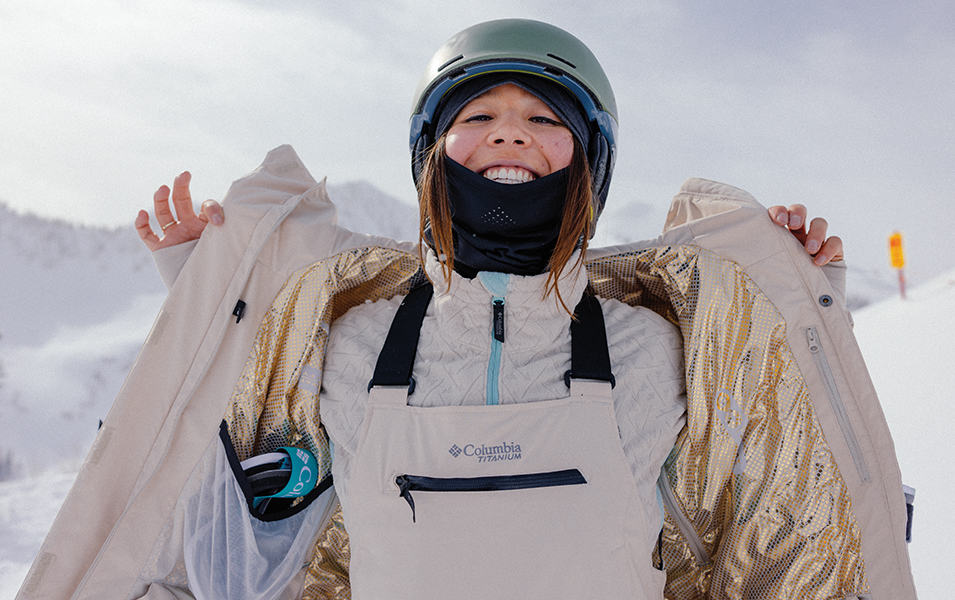 A skier wearing a white Columbia Sportswear jacket smiles at the camera as she peels off her layers. 