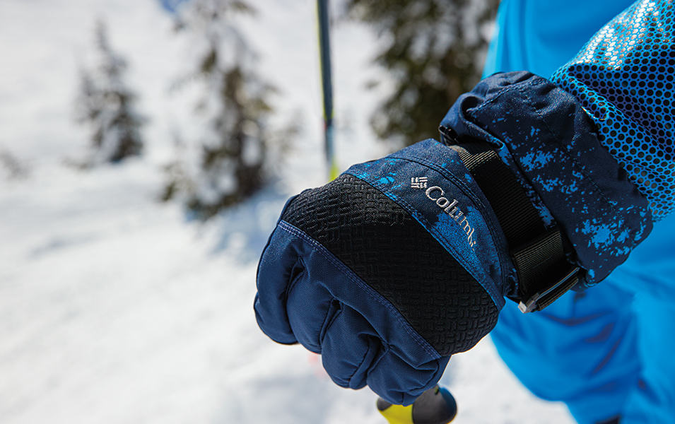 A close-up shot of a blue Columbia Sportswear glove gripping a ski pole in a snowy mountain setting. 